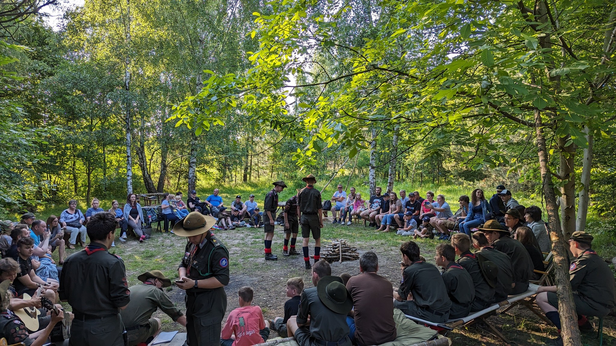 Harcerze oraz ich rodzice zgromadzenie dookoła ogniska. Trzech harcerzy przy ognisku czeka aby je rozpalić. Dookoła widać drzewa. Ognisko jest trochę wkopane w ziemię
